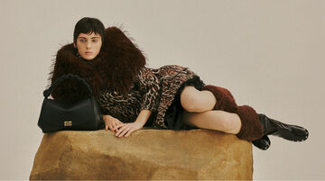 An editorial campaign photo of a person wearing a patterned dress with a large textured brown fur collar, reclining on a large stone block next to a black handbag