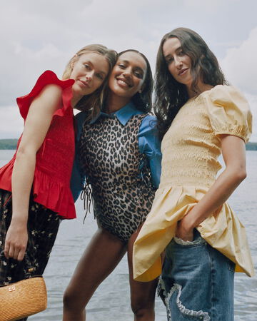 Three models standing together by a lake wearing summer outfits including a red ruffled smocked top, a leopard print vest over a blue shirt, and a yellow puff-sleeve top