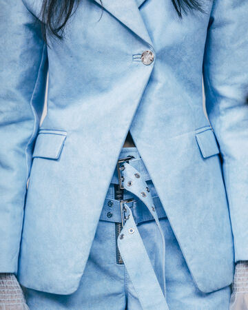 Close-up of light blue tailored blazer fabric layers detailed with metallic eyelets, double silver buckles, and a heavy metal zipper