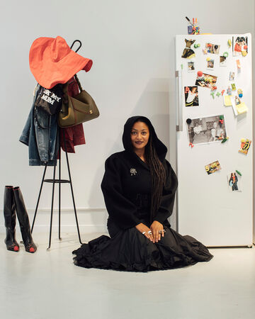 Model in a black hooded coat and voluminous black skirt sitting on the floor next to a coat rack and a photo-covered white refrigerator