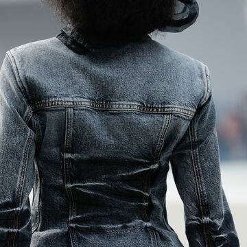 Back view close-up of a structured grey denim or woven jacket featuring distinct vertical seams and lacing details