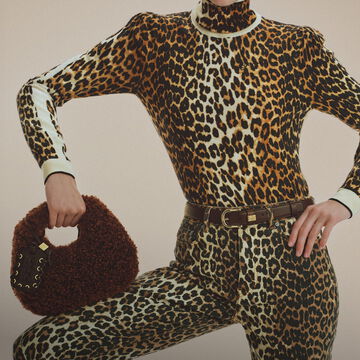 Model wearing a matching leopard-print turtleneck sweater and leopard-print jeans belted at the waist, holding a brown shearling mini handbag.