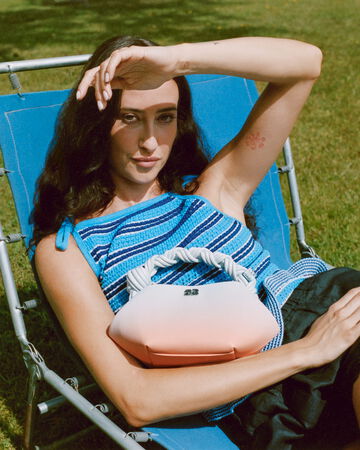 A model lounging outdoors in a lawn chair wearing a blue striped crochet top, holding a light pink to coral gradient GANNI Bou Bag