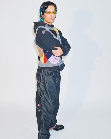 Sahar Jamili design look: A person wearing a dark hooded utility zip-up jacket decorated with colorful woven panels and loose black cargo pants