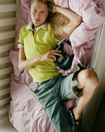Model lying on pink bedsheets wearing a lime green polo shirt, denim Bermuda shorts, and holding a black GANNI shoulder bag