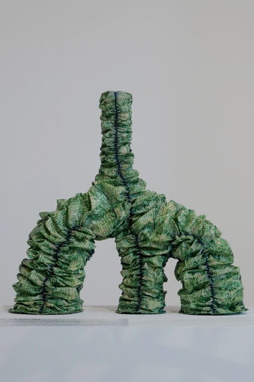 An abstract, three-legged organic sculpture crafted entirely from textured green plant-based material tiles, displayed on a white pedestal