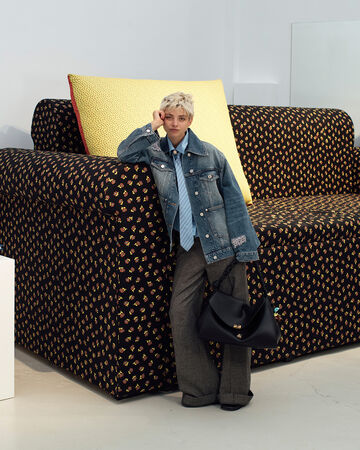 Model with short blonde hair wearing a denim jacket, tie, and trousers holding a black shoulder bag while leaning against a large patterned couch