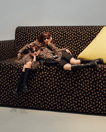 Two models in matching animal-print outfits and sunglasses lounging together on an oversized patterned sofa