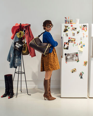 Model wearing a blue cardigan, animal-print skirt, and brown knee-high boots holding a large dark shoulder bag next to a white refrigerator covered in photos