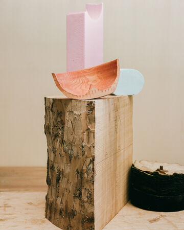 A modernist sculptural arrangement featuring a terracotta curved piece balancing on a wooden pillar alongside light blue and pink geometric blocks