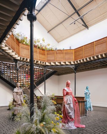GANNI Spring Summer presentation display featuring mannequins dressed in layered floral and patterned sheer gowns arranged around a wooden staircase with large wildflower installations