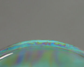 Macro close-up view highlighting the curved, iridescent edge of a smooth surface reflecting a spectrum of rainbow colors against a neutral grey backdrop