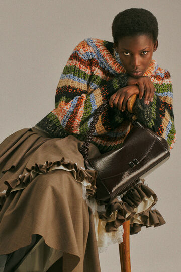 An editorial campaign photo of a person resting their hands and chin on the back of a wooden chair, wearing a thick, textured knit sweater with multi-colored horizontal stripes