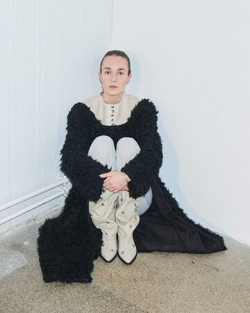 Nicklas Skovgaard design garment: A person sitting in a corner wearing an oversized black textured, ruffled winter coat with white circular collars