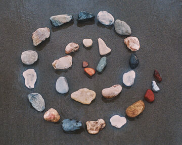 A loose circular arrangement of multi-colored, smooth natural stones scattered on a textured dark grey surface