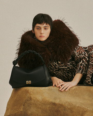 Model reclining on a stone platform wearing a patterned outfit with oversized fur collar and black handbag