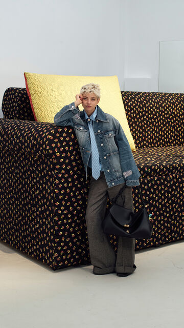 Model with short blonde hair wearing a denim jacket, tie, and trousers holding a black shoulder bag while leaning against a large patterned couch