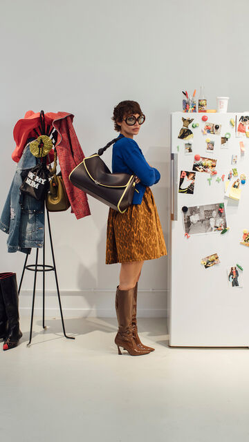 Model wearing a blue cardigan, animal-print skirt, and brown knee-high boots holding a large dark shoulder bag next to a white refrigerator covered in photos